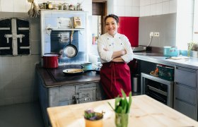 Kräuterwirtin Gerda Stocker neben ihrem fast 100 Jahre alten Holzofen, © Nicole Heiling Eine Köchin in weißer Jacke und roter Schürze steht in einer Küche vor einem alten Herd.