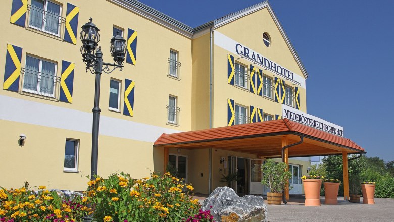 Hochzeitspavillon, © Besta Betriebs GmbH Außenansicht eines gelben Hotels mit blauen Fensterläden und einem Vordach. Vor dem Gebäude blühen gelbe und pinke Blumen.