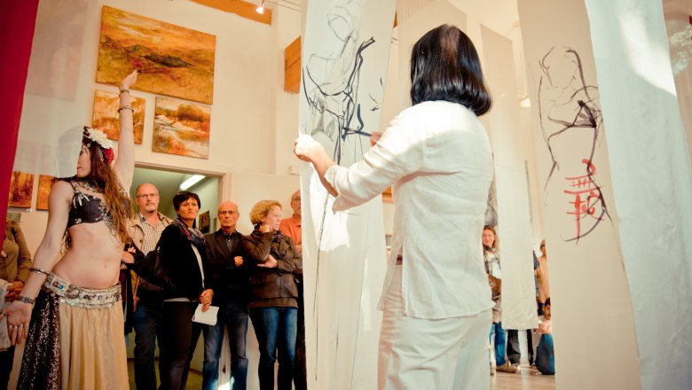 Atelier Buchner, © Christine Buchner, Foto Birgit Muehleder Eine Frau in weißer Kleidung betrachtet Zeichnungen auf hängenden Stoffbahnen in einer Galerie mit Zuschauern und Gemälden an der Wand.