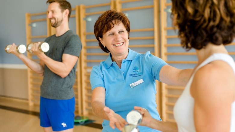 Gesundheitswoche, © Niederösterreich-Werbung/Doris Schwarz-König Eine Trainerin in einem blauen Polohemd hilft einer Frau beim Hanteltraining, während ein Mann im Hintergrund ebenfalls trainiert.