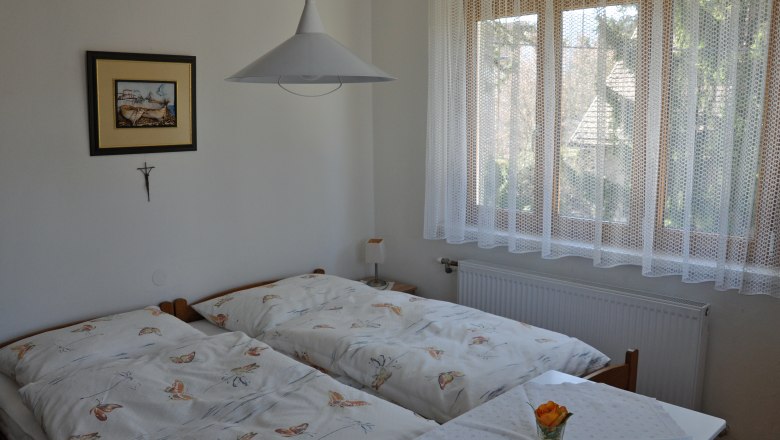 Gemütliche Zimmer bei Familie Kremsl, © Familie Kremsl Gemütliches Schlafzimmer mit zwei Einzelbetten, einem Bild an der Wand und einem Fenster mit Spitzenvorhang.