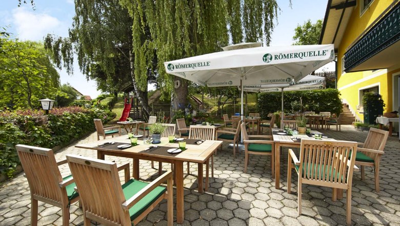 Gastgarten, © Wiener Alpen / Bene Croy Ein sonniger Gastgarten mit Holzmöbeln und Sonnenschirmen, umgeben von grüner Vegetation und einem gelben Gebäude.