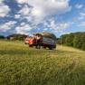 Field work, © Wachahof A tractor with a trailer full of hay drives across a green field under a blue sky with clouds.