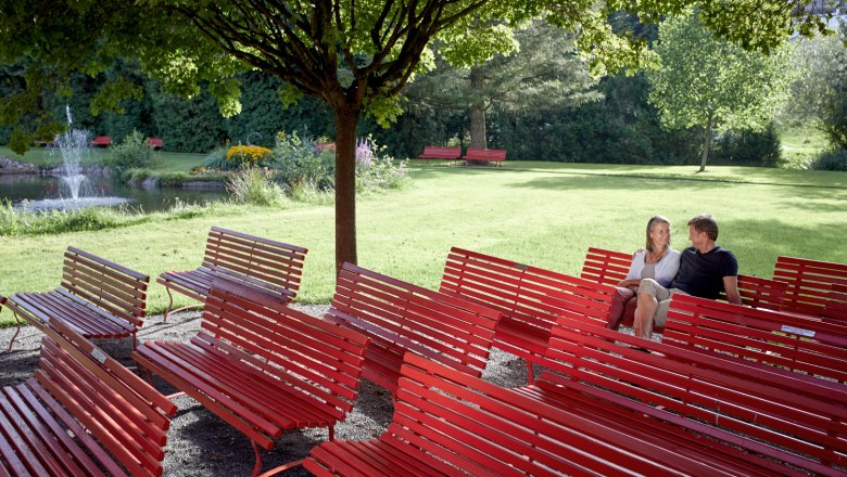 Music pavilion Kurpark, © Wiener Alpen, Florian Lierzer A couple sits on red benches in the spa gardens, surrounded by trees and a pond with a fountain.
