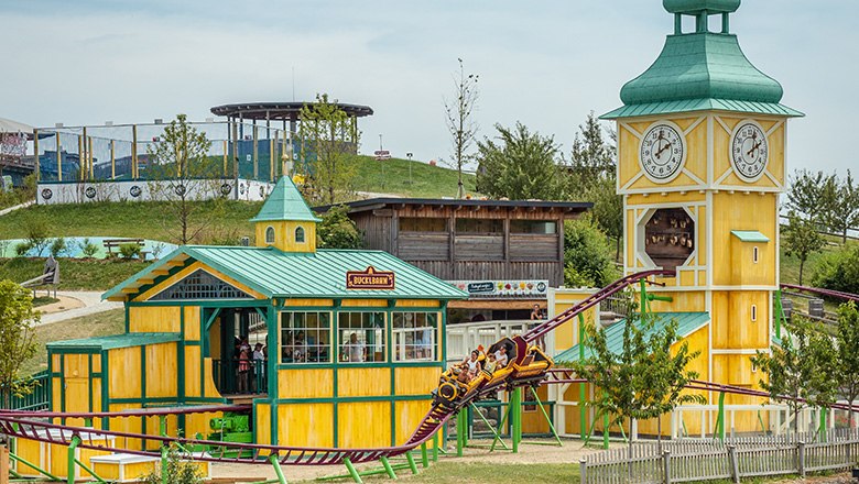 Eis-Greissler adventure park Bucklbahn, © Blochberger Eisproduktion GmbH The "Bucklbahn" roller coaster in the Eis-Greissler Adventure Park with yellow buildings and a green hill in the background.