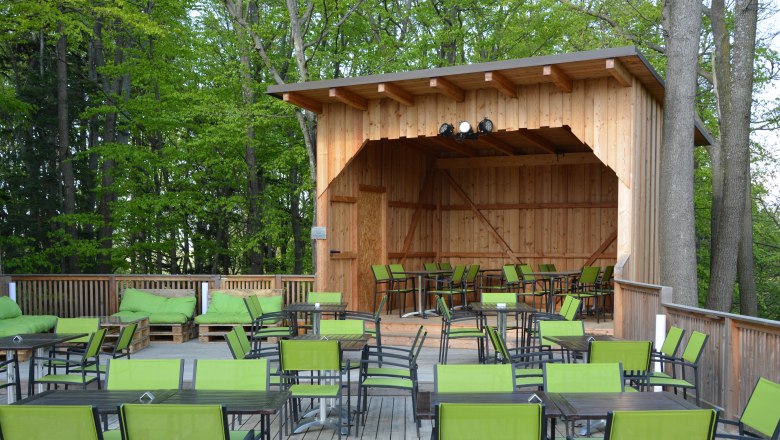 Gemütlicher Terassenbereich im Turmcafé Schwarzenbach, © Gemeinde Schwarzenbach Eine Holzterasse mit schwarzen Tischen und grünen Stühlen und einer überdachten Holzbühne.