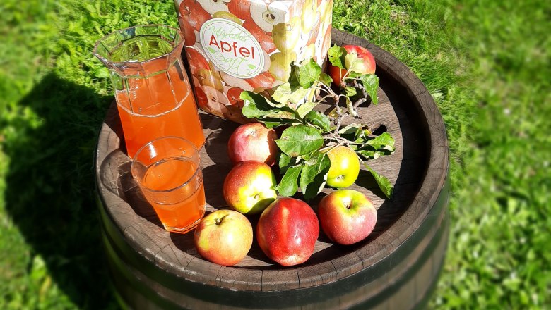 Organic apple juice, © Biohof Puchegger A jug and a glass of naturally cloudy apple juice and fresh apples on a wooden barrel standing on a green meadow.