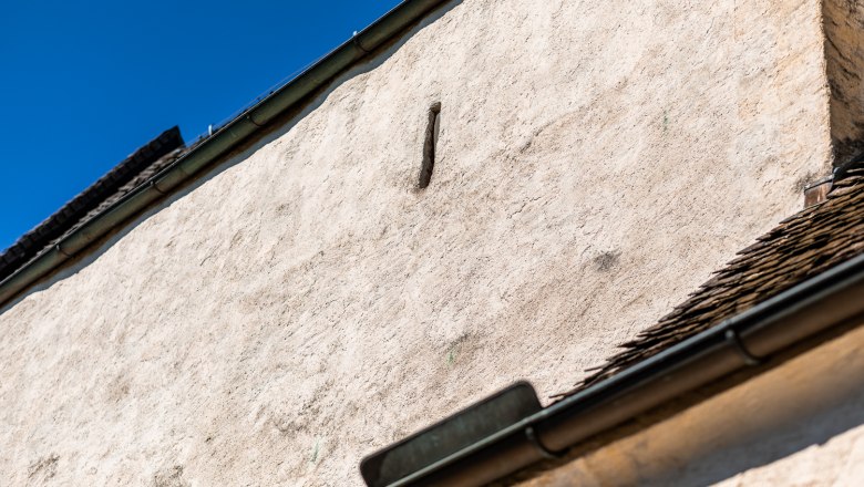 Wehrkirche Bad Schönau, © Wiener Alpen, Christian Kremsl Nahaufnahme einer weißen Mauer mit Schießscharte Dachrinne und blauen Himmel im Hintergrund.