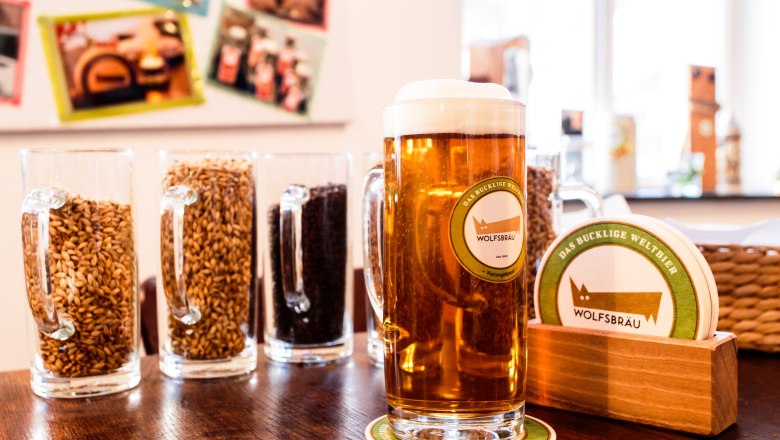 Wolfsbräu - the humpbacked world beer, © Familie Wagner Mug with beer next to beer mats, in the background beer glasses with grain.
