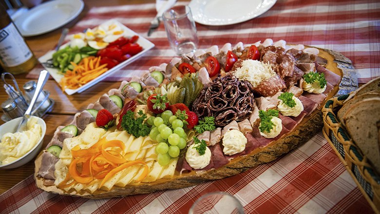 Pleasure farm, © XXXXX A rich snack on a wooden plate with sausage, cheese, vegetables and bread on a red and white checkered tablecloth.