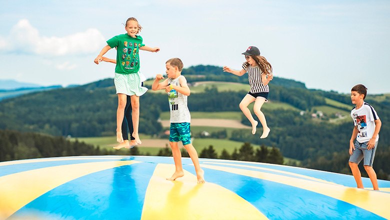 Eis-Greissler adventure park bouncy cushion, © Blochberger Eisproduktion GmbH Four children jump on a colorful bouncy cushion in front of a hilly landscape with lots of trees.
