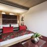 Zimmer Magnolie_Orchidee Linsberg Asia, © Linsberg Asia Modernes Hotelzimmer mit Doppelbett, roten Kissen und Blumenmuster. Ein Glas-Couchtisch mit einer Vase voller Tulpen steht im Vordergrund.