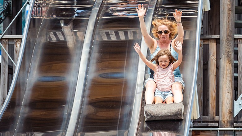 Eis-Greissler adventure park slide tower, © Blochberger Eisproduktion GmbH A woman and a child with raised arms sit sliding on a mat on a silver slide.