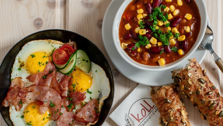 Warme Snacks, © Koll GmbH Verschiedene warme Snacks auf einem Tisch, darunter Spiegelei mit Speck, Sandwiches und eine Schüssel Chili.