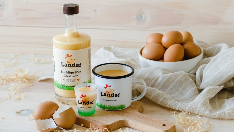 Eierlikör, © Vikografik/ Viktoria Kornfeld Eine Flasche, ein Glas und eine Tasse gefüllt mit Eierlikör auf einem Holzbrett. Im Hintergrund eine Schale mit Eiern auf einem grauen Tuch.