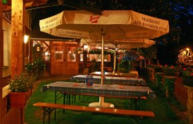 Krenn Wirt, © Krenn Wirt Ein gemütlicher Biergarten bei Nacht mit Holztischen, blau karierten Tischdecken und großen Sonnenschirmen der Marke Stiegl.