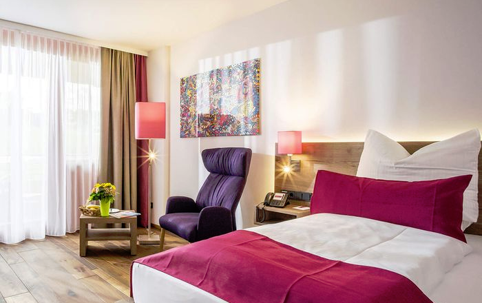 Zimmer, © Vivea Gesundheitshotel Gemütliches Zimmer mit Doppelbett, Ohrensessel, Tisch, Lampen, einem Wandbild und Holzboden.