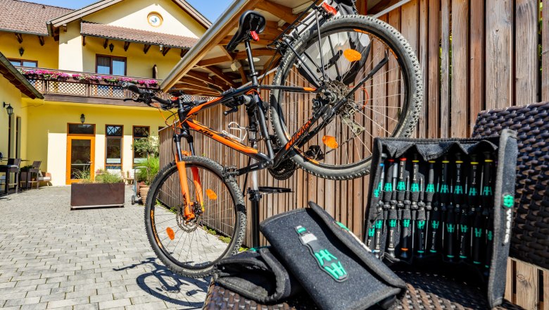 Radgastgeber Hotel Post-Hönigwirt, © Wiener Alpen/Martin Fülöp Ein orangenes Fahrrad auf einem Ständer vor einem gelben Gebäude mit Holzzaun und Werkzeugset im Vordergrund.