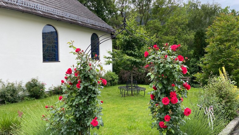 Rose arch by the chapel opposite the inn, © Wiener Alpen A rose arch with red roses in front of a chapel with colorful windows and a green garden.
