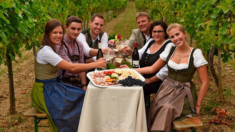Familie Fingerlos, © Fingerlos Eine Familie sitzt auf einem Tisch mit Wein, Wurstplatten und Weingläsern zwischen Weinstöcken.