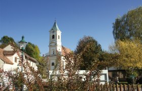 Pfarrkirche Walpersbach, © Roffeis Heinz Pfarrkirche Walpersbach mit Turm und umliegenden Gebäuden, umgeben von Bäumen und Sträuchern unter blauem Himmel.