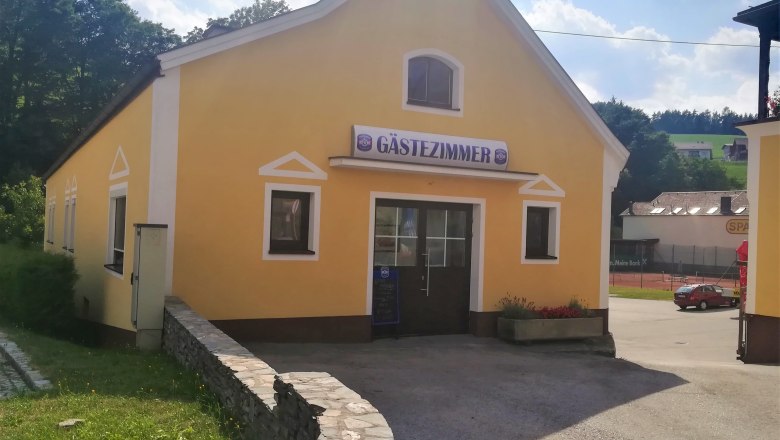 Gästezimmer beim Gasthof Gansterer, © Wiener Alpen Gelbes Gebäude mit Schild 'Gästezimmer', umgeben von Bäumen und einem Parkplatz.