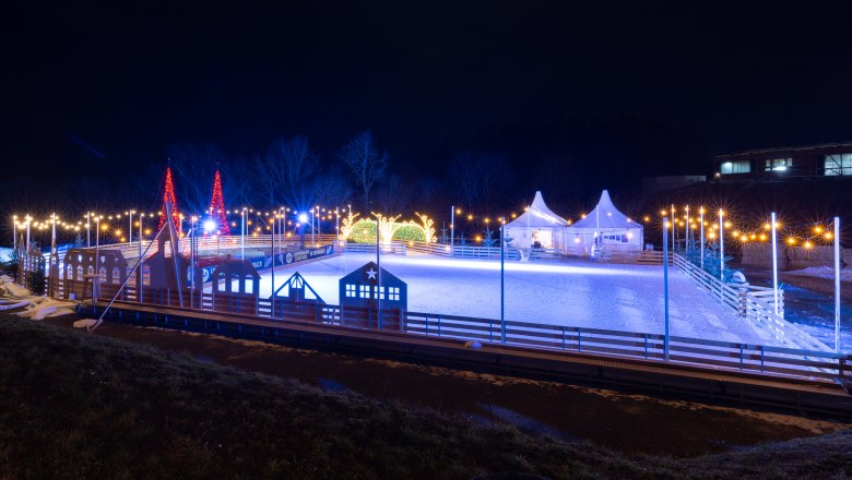 Ice skating rink in the Eis-Greissler Welt, © Blochberger Eisproduktion GmbH Illuminated ice rink at night with decorative lights and tents.