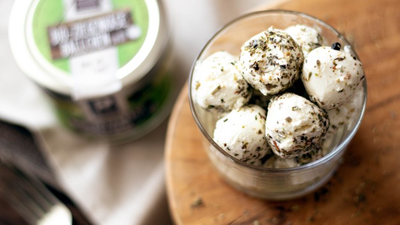 Goat's cheese balls, © Sooo gut schmeckt die Bucklige Welt/Viktoria Kornfeld A glass filled with goat's cheese balls, sprinkled with herbs, on a round wooden board. In the background, another jar of goat's cheese balls with a green label.