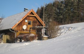 Wachahof im Winter, © Wachahof Ein verschneites Holzhaus am Waldrand mit blauem Himmel.