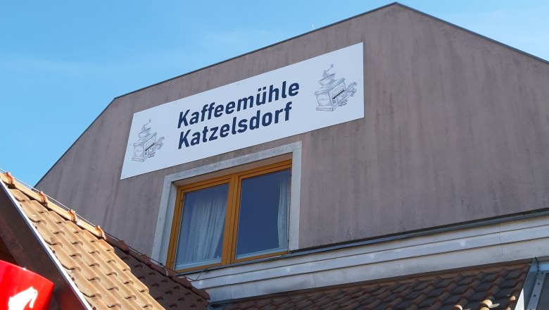Kaffeemühle, © Kaffeemühle Katzelsdorf Komböck Gebäude mit Schild 'Kaffeemühle Katzelsdorf' und Zeichnung einer Kaffeemühle.