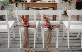 Tailor's goods, © Julia Geiter, Angelika Riegler White chairs decorated with flowers and fabric on a wooden terrace, prepared for a wedding.