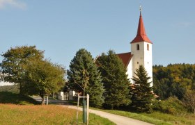 Pfarrkirche Ofenbach, © Thermengemeinden Pfarrkirche Ofenbach mit rotem Turmdach, umgeben von Bäumen und Wiesen.