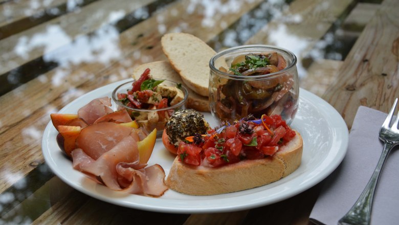 Schlemmen im Turmcafé, © dasTurmcafé - Gemeinde Schwwarzenbach Ein Teller mit Brot, Schinken, Antipasti, Bruschetta und Obst auf einem Holztisch.