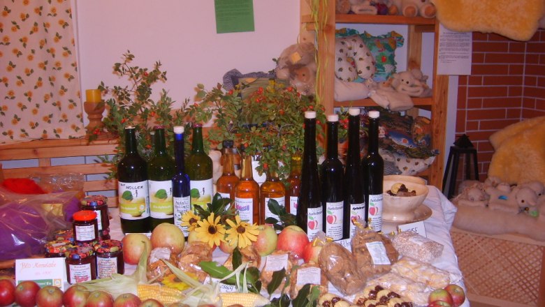 Hofladen im Bio-Schafbauernhof Kölbel, © Kölbel Ein Tisch in einem Hofladen mit verschiedenen Bio-Produkten wie Äpfeln, Marmeladen, Säften und Likören, dekoriert mit Blumen und Pflanzen.