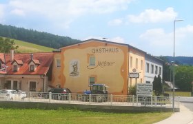 Café Restaurant Mößner in Schwarzenbach, © Wiener Alpen Außenansicht des Gasthauses Mößner, gelb-orange gestrichen, in einer ländlichen Umgebung mit blauer Traktor und Autos vor dem Gebäude.