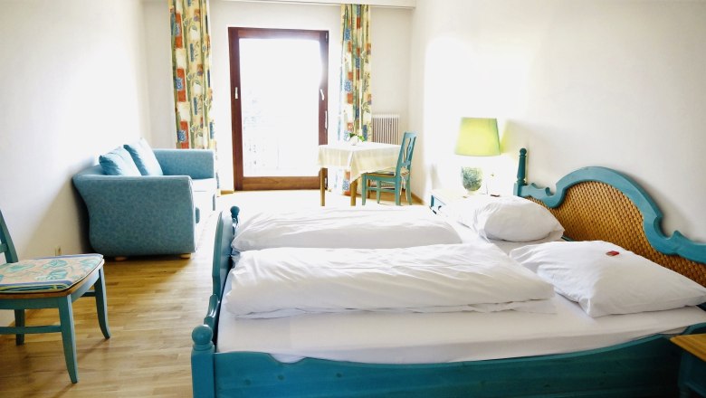 Großes Zimmer, © Seminarhotel NaturSinne Ein helles Zimmer mit einem Doppelbett, blauer Couch, Tisch und Stuhl vor einem Fenster mit Vorhängen.