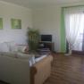Living room, © Anke Spreitzer A living room with a white sofa, a small TV, plants and large windows with curtains.