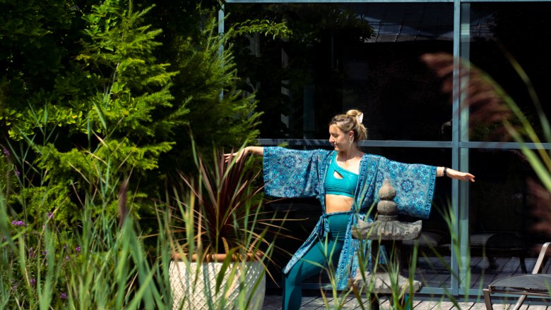 Linsberg Asia - Ein Tag voller Leichtigkeit, © Linsberg Asia Eine Frau in blauer Yogakleidung macht eine Yoga-Pose im Freien neben einem Teich und Pflanzen.