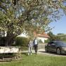 E-mobility, © Wiener Alpen A couple walks next to a parked electric car under a blossoming tree in a rural garden.