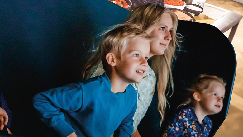 Eis-Greissler ice cream time travel & flying theater, © Blochberger Eisproduktion GmbH Two children and a woman sit in a carriage in the Flying Theater and look ahead, spellbound.