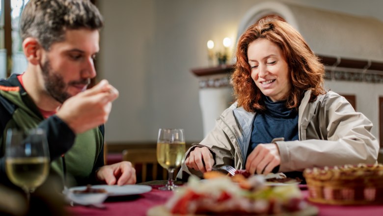 At the Böhm gourmet farm and wine tavern in Katzelsdorf, © Wiener Alpen/Fülöp, Kremsl A man and a woman sit in a parlor on a rustic wooden table and eat a Brettljause snack