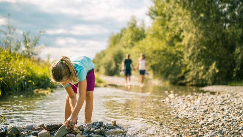 A child plays in a shallow stream while two people walk in the background.