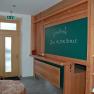 Innenansicht eines Hotels mit einer Tafel, auf der 'Gasthof Zur Alten Schule' steht, und einem Regal mit Broschüren.