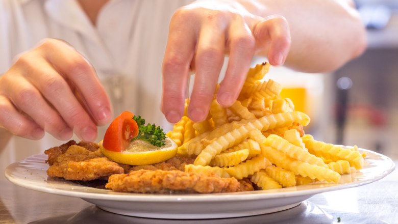 Ein Teller mit Wiener Schnitzel, Pommes frites, Zitrone und Tomate wird von einer Person angerichtet.