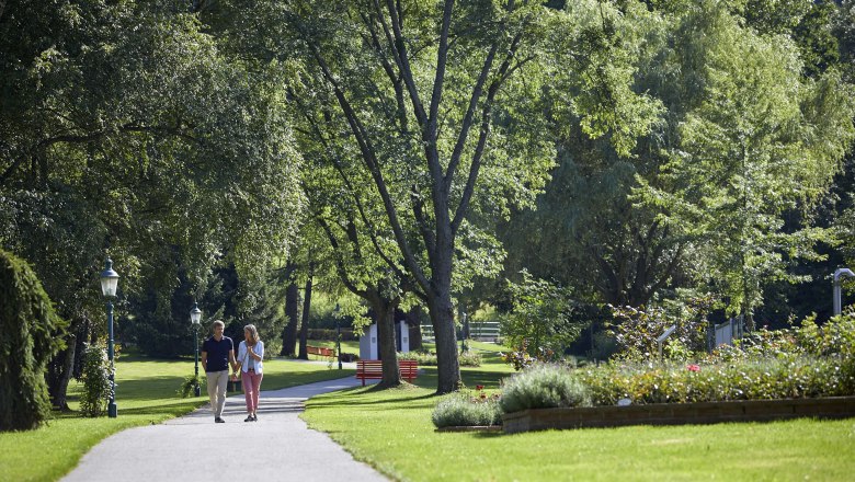 A couple walks along a path in the spa gardens of Bad Schönau, surrounded by green trees and well-tended flowerbeds.