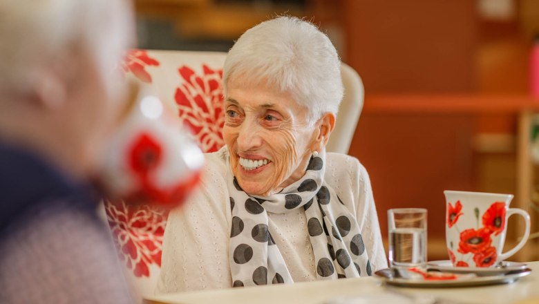 Elderly woman sitting at a table smiling, in the foreground a cup with a floral pattern and a glass of water.