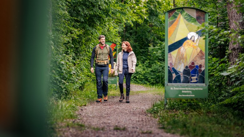 Zwei Personen wandern auf einem Waldweg mit gläsernen Kunstwerken am Rand.