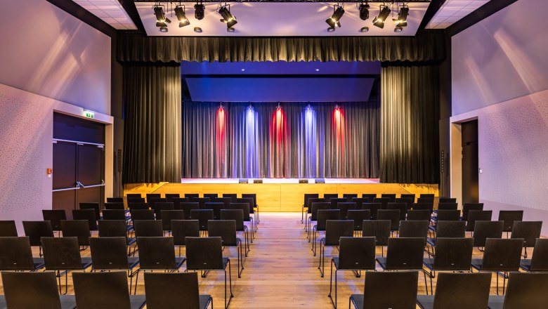 Ein leerer Veranstaltungssaal mit Stuhlreihen und einer B&uuml;hne mit Vorh&auml;ngen und bunter Beleuchtung.