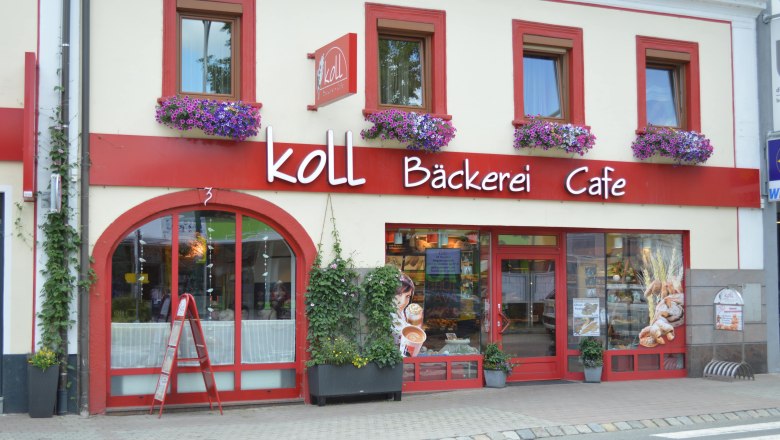 Außenansicht der Koll Bäckerei Café in Kirchschlag mit roten Fensterrahmen und Blumenkästen.