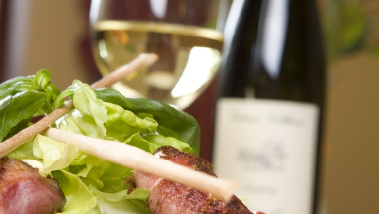 Ein Teller mit Salat und Käse im Speckmantel, im Hintergrund ein Weinglas und eine Weinflasche.
